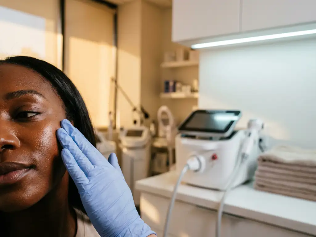 Huidanalyse van een donkere vrouwenhuid door behandelaar met handschoen, modern esthetisch kliniekinterieur met medisch apparaat op tafel.