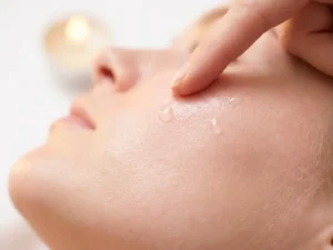 Vingertop die hydraterende serum aanbrengt op stralende huid, druppels zichtbaar op het huidoppervlak, witte achtergrond.