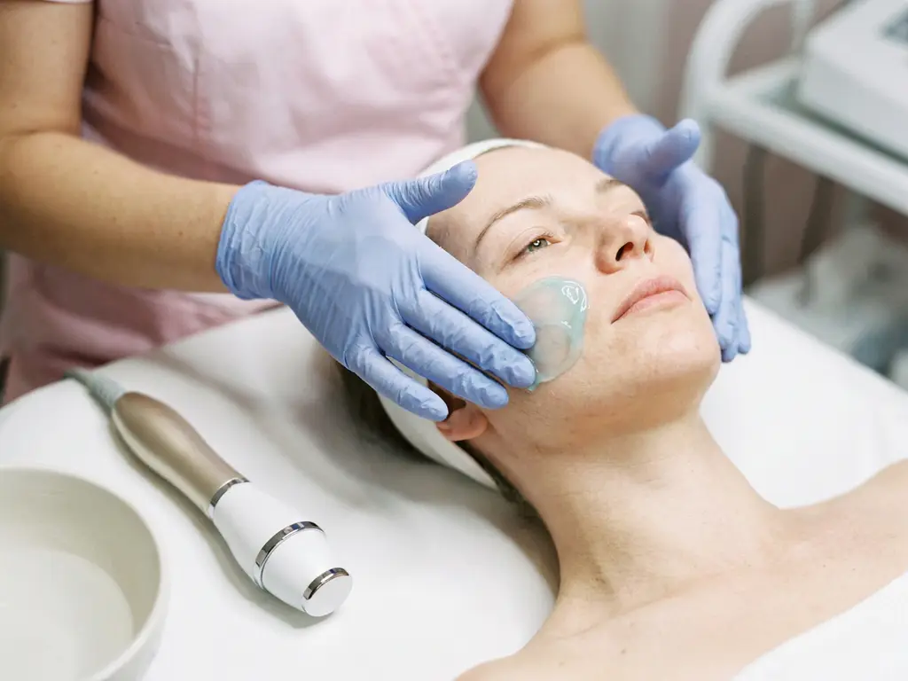 Estheticienne die hydraterende gel aanbrengt op een gevoelige huid, klinisch apparaat op witte behandeltafel op de achtergrond.