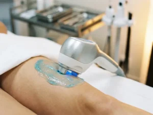 Shockwave therapie handstuk op dijbeen met blauw geleidingsgel, klinische behandeltafel op de achtergrond.
