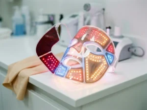 LED-gezichtsmasker met rood, blauw en amber licht op wit klinisch oppervlak, omgeven door zacht uitstralend licht.