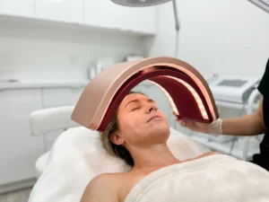 Vrouw ondergaat LED-lichttherapie op haar gezicht met rozegouden en rode lichtpanelen in een minimalistische witte kliniek.