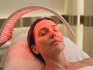 Vrouwengezicht tijdens roodlichttherapie in een klinische omgeving, zachte karmozijnrode gloed verlicht een gladde huid met fijne lijntjes.